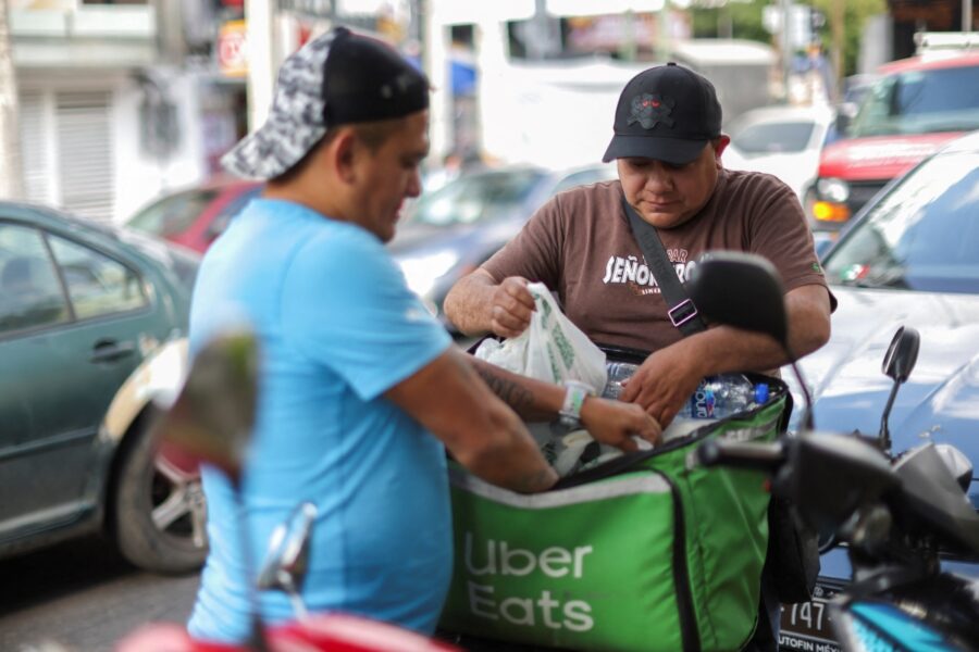 Dois entregadores de Uber Eats interagindo na rua, um deles está colocando itens na bolsa de entrega verde, enquanto o outro observa atentamente.