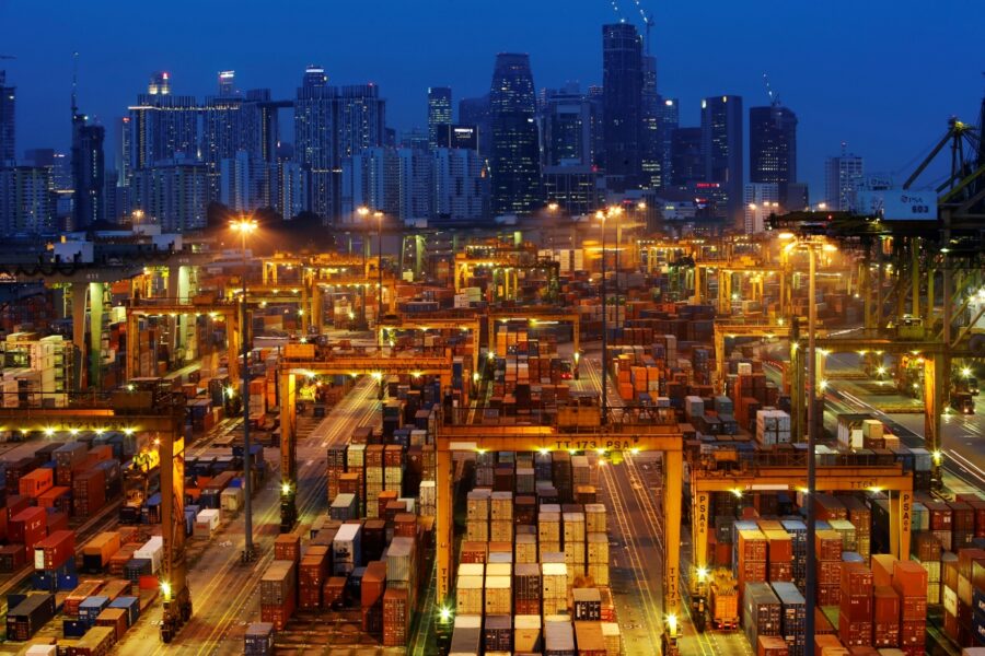 Vista noturna de um terminal portuário moderno repleto de contêineres, com os arranha-céus de uma cidade ao fundo, destacando a movimentação no comércio internacional.