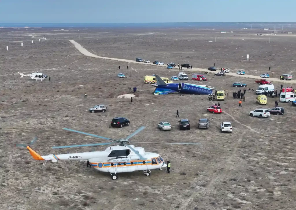 Vista aérea de um helicóptero e veículos de emergência em um local de acidente, incluindo um avião pousado em uma área desértica.