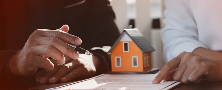 Imagem de mãos de pessoas assinando documentos de compra de imóvel, com uma maquete de casa ao fundo, simbolizando a importância do registro eletrônico de imóveis