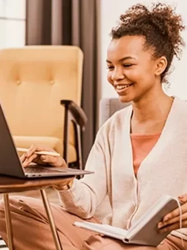 Mulher sorridente trabalhando em casa, sentada no chão com laptop, caderno e caneca, ambiente aconchegante e moderno.