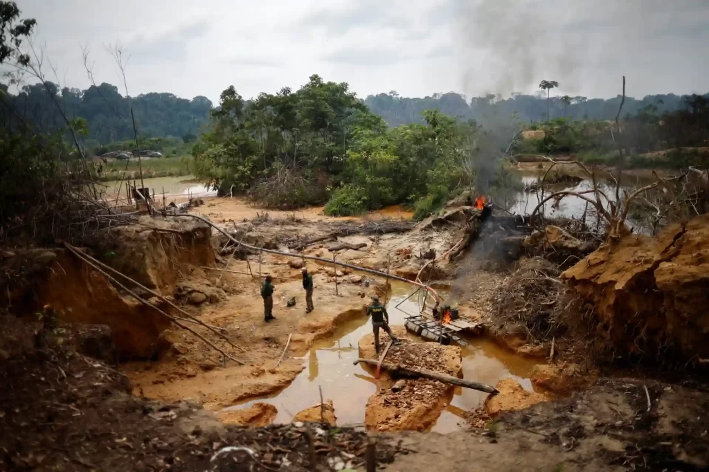 Imagem de uma área devastada na Amazônia, com homens trabalhando na extração de ouro e fumaça de incêndios ao fundo. A cena ilustra os impactos da mineração ilegal e desmatamento na floresta.