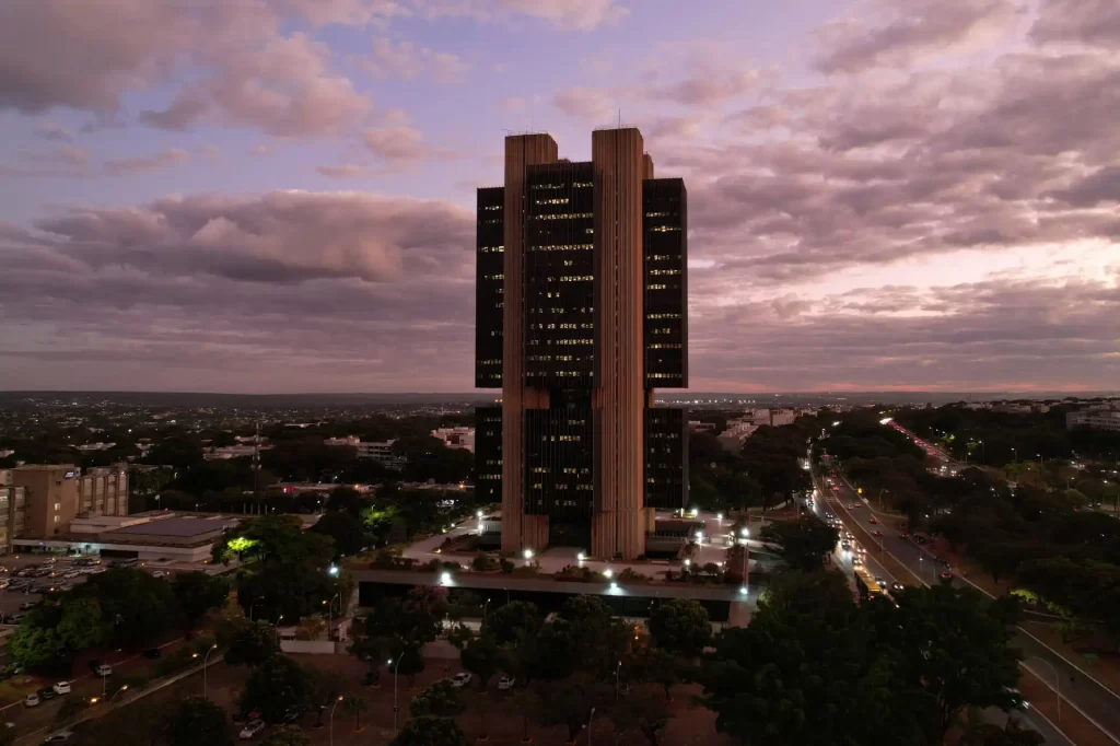 Imagem do edifício da sede do Banco Central do Brasil iluminado ao entardecer, destacando sua arquitetura moderna e imponente a céu aberto.