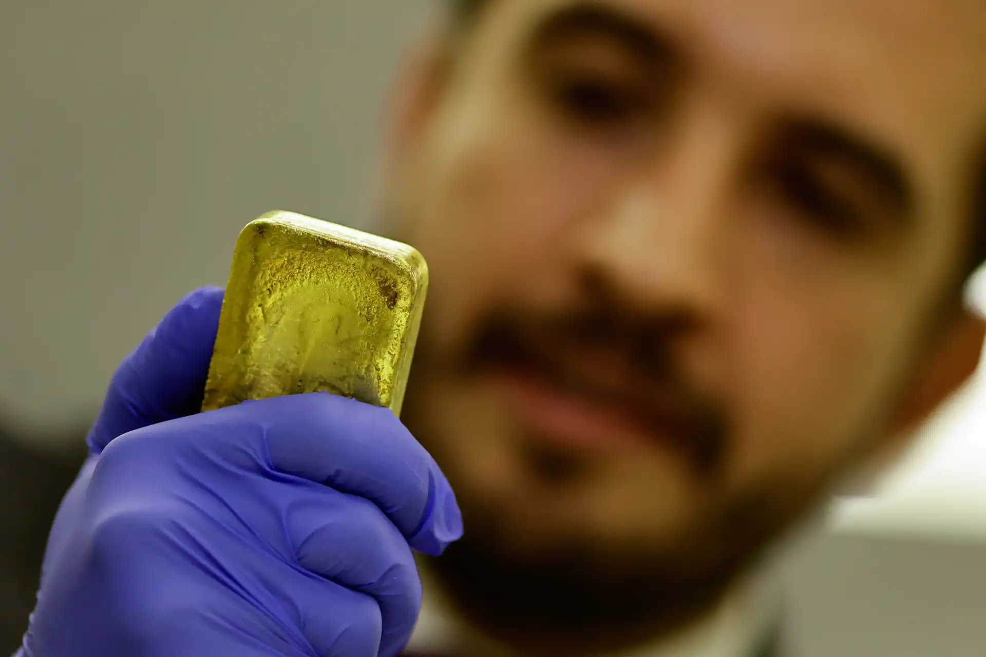 Homem usando luvas azuis segurando uma barra de ouro. Foco em seu olhar e na barra reluzente, destacando a importância do ouro como investimento e símbolo de riqueza.