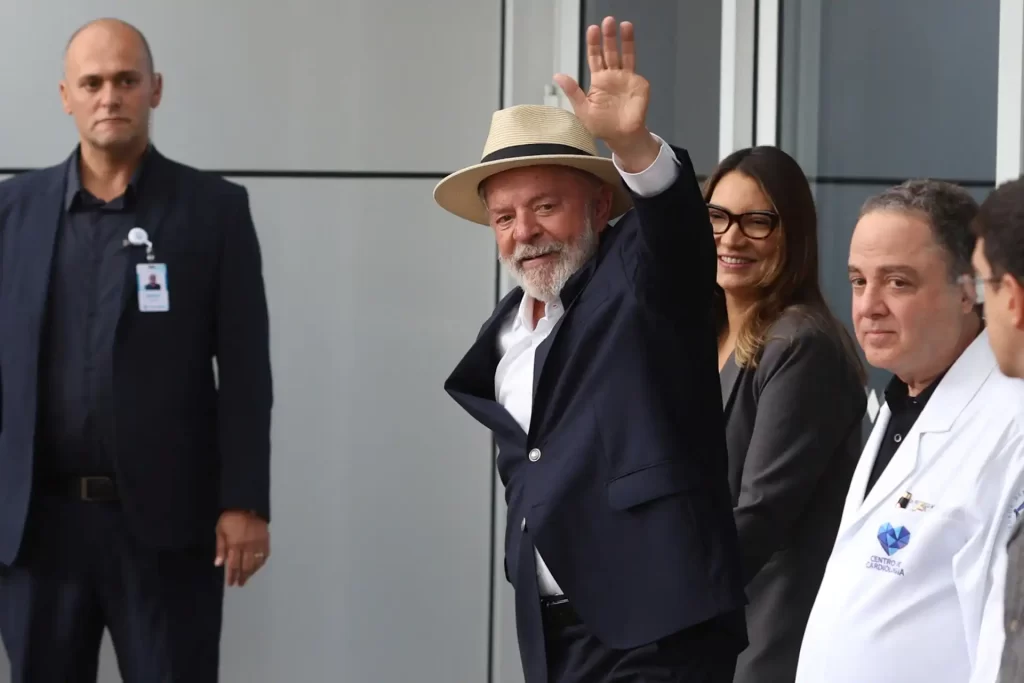 O presidente Luiz Inácio Lula da Silva acena em meio a assessores, vestindo terno e chapéu, em evento recente, demonstrando simpatia e carisma.