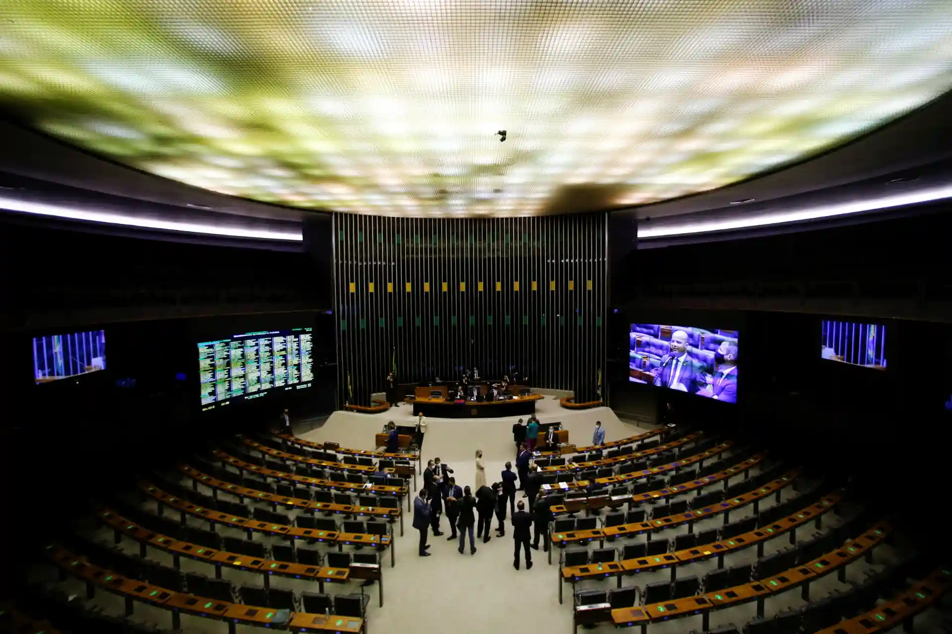 Vista interna do Plenário da Câmara dos Deputados em Brasília, com deputados em atividade e painéis eletrônicos ao fundo.