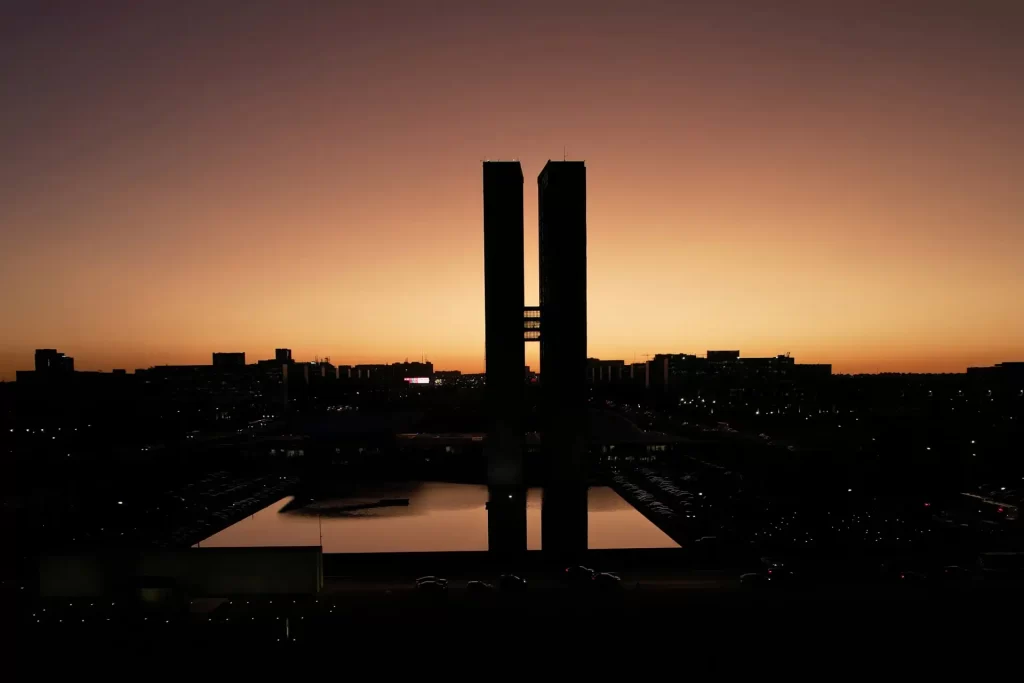 Imagem do pôr do sol com silhuetas de torres na cidade, refletindo em um lago. Um cenário urbano impressionante que captura a beleza do crepúsculo.