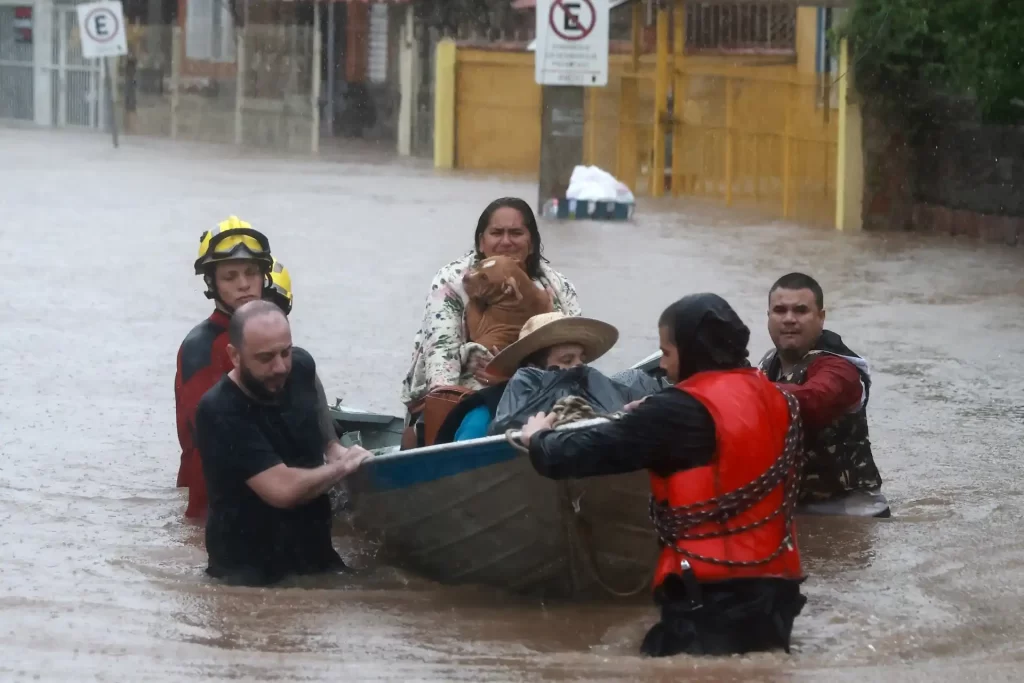 Imagem de um resgate durante enchente, com pessoas em uma canoa sendo ajudadas por bombeiros em área inundada.