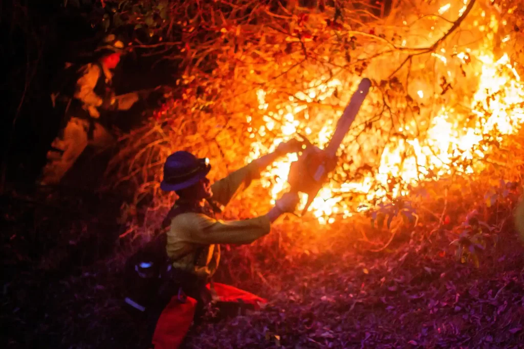 Bombeiros combatendo incêndio florestal à noite, utilizando motosserra para cortar a vegetação em chamas, com chamas iluminando a cena ao fundo.