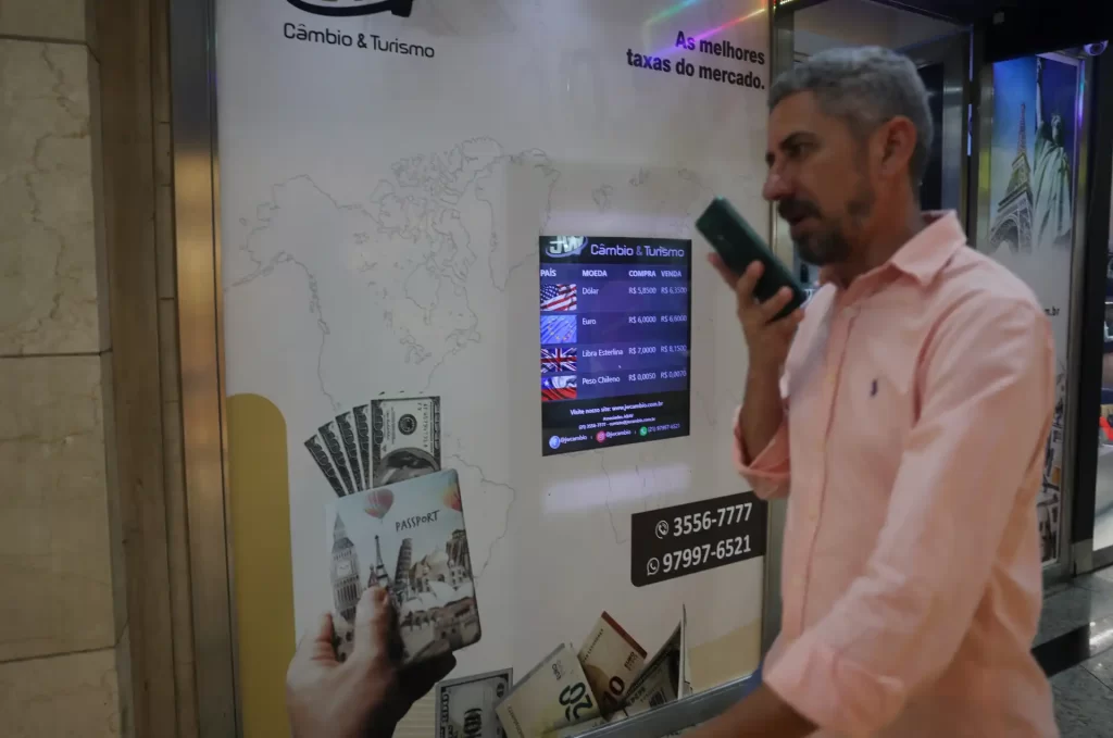 Homem utilizando um telefone próximo a um painel de câmbio e turismo, com notas de dinheiro na mão e informações de taxas de câmbio visíveis.