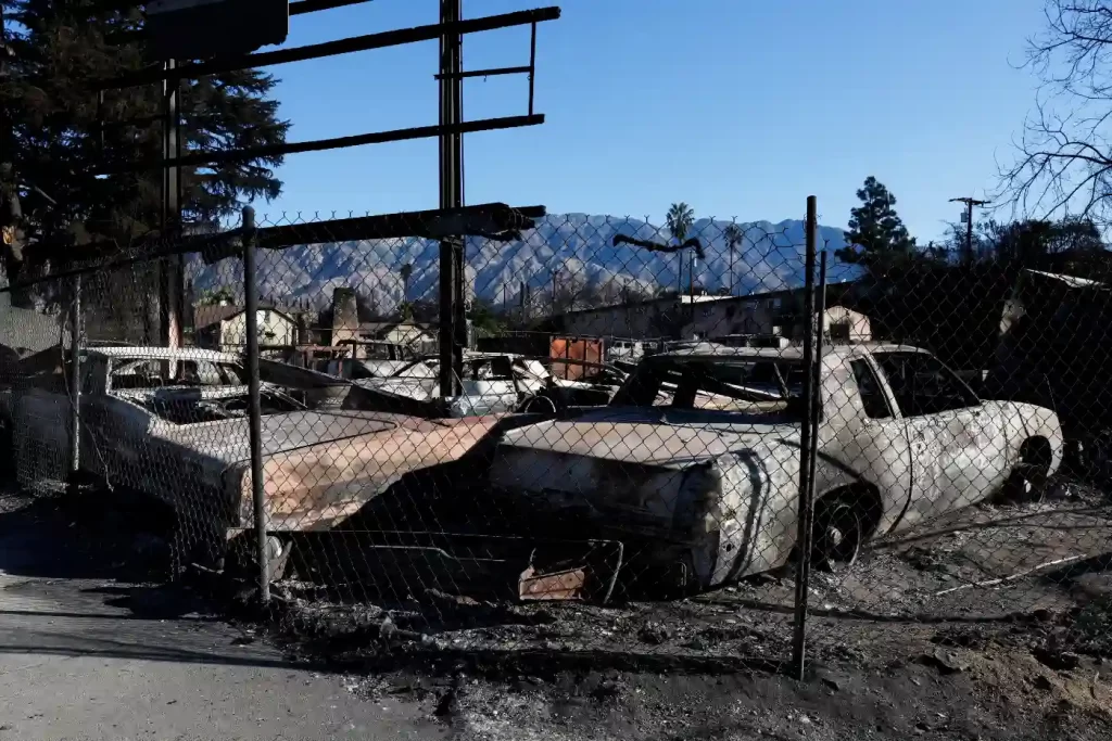 Imagem de carros queimados e danificados em uma área devastada, com montanhas ao fundo e cercas ao redor, evidenciando os danos de um incêndio.