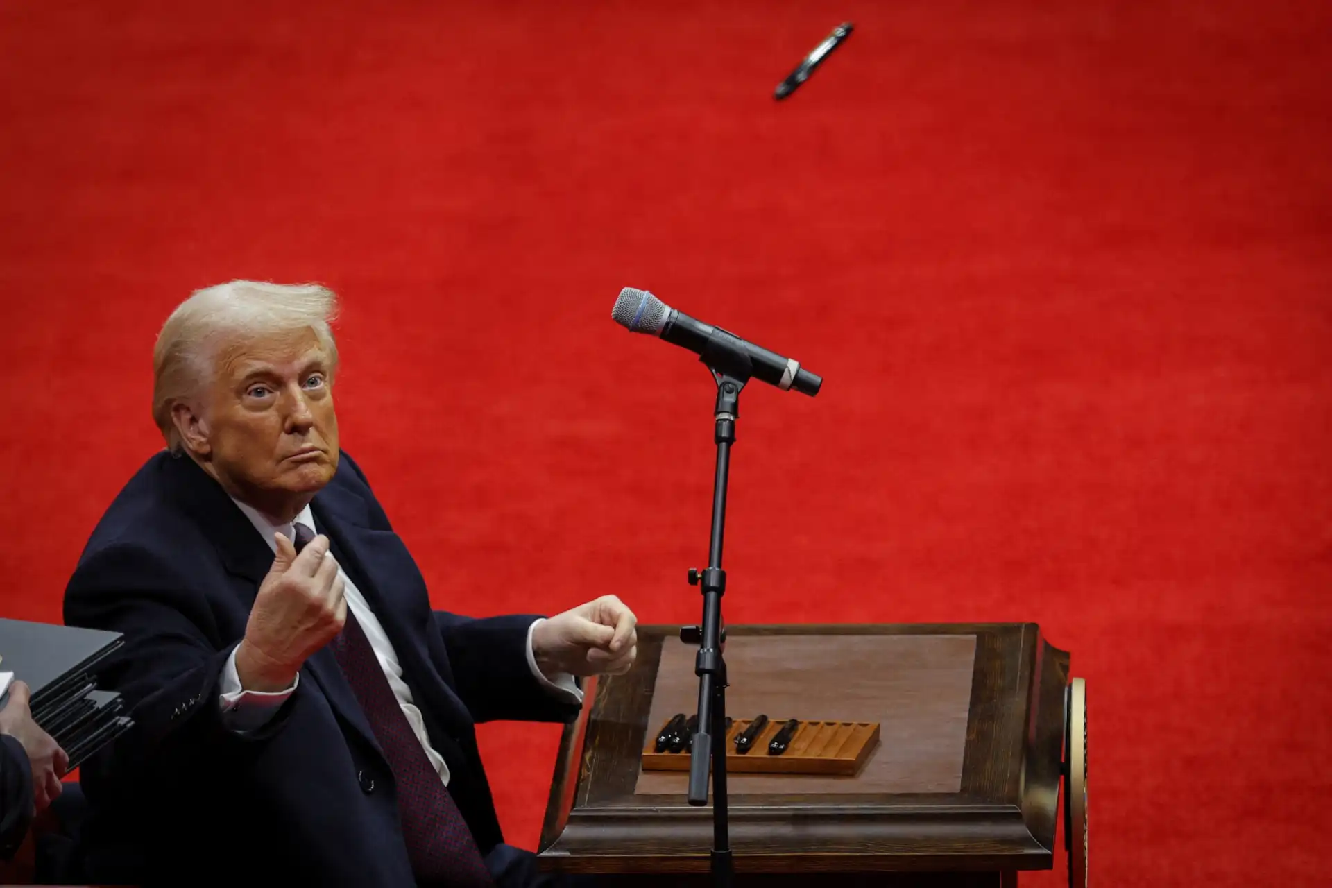 Imagem de Donald Trump em um evento, olhando para a câmera com expressão séria, destacando o fundo vermelho e um microfone ao seu lado.