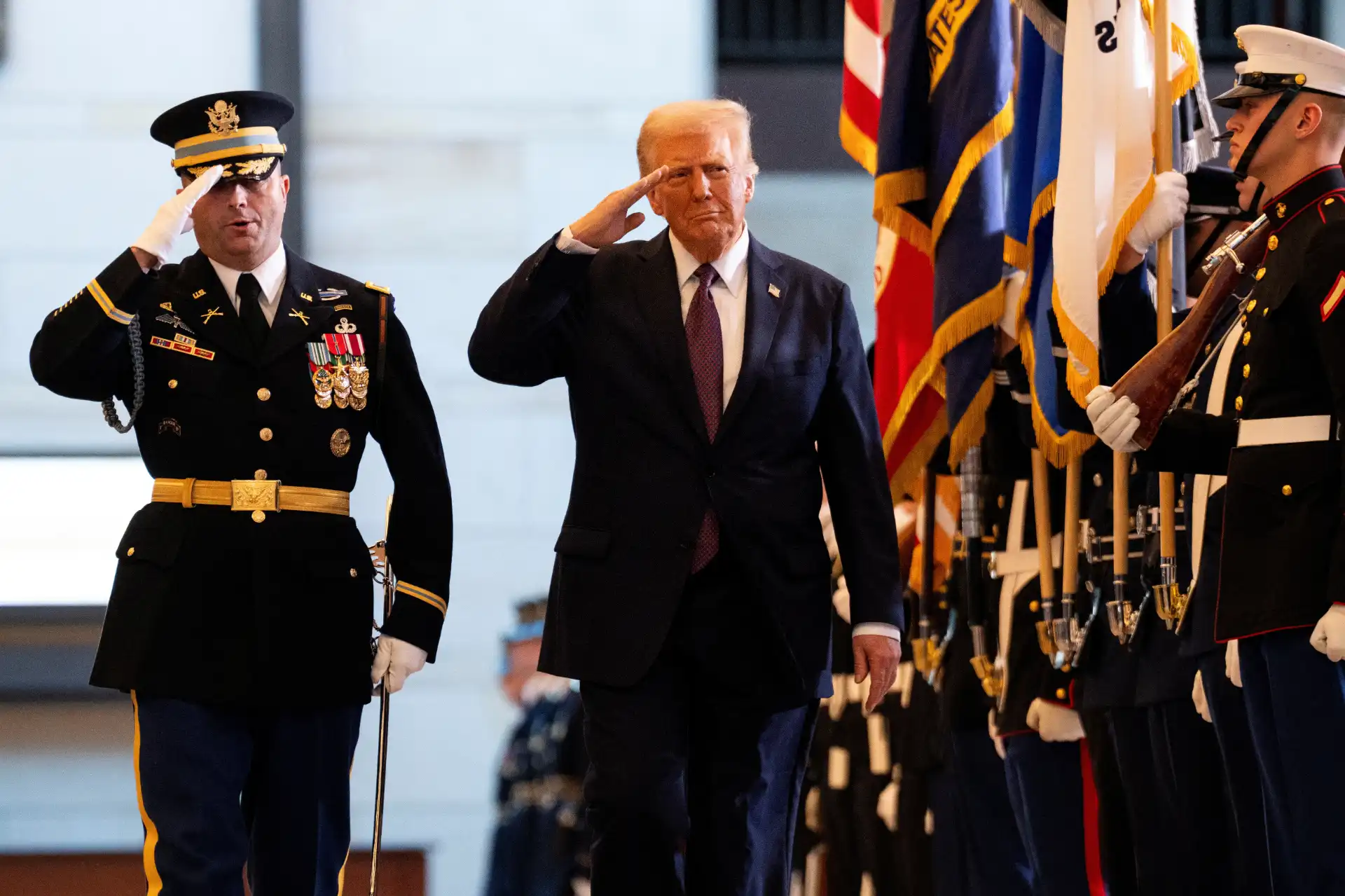 Donald Trump prestando homenagem ao lado de um oficial militar durante cerimônia oficial, com bandeiras ao fundo