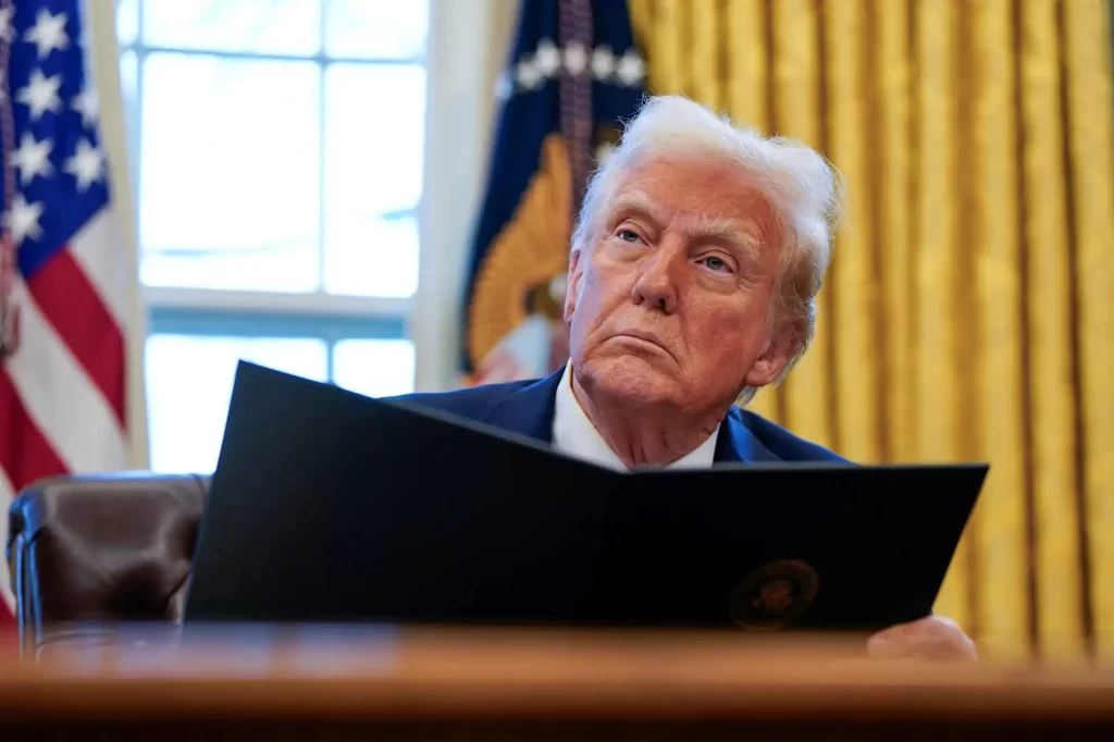 Imagem de Donald Trump sentado no Salão Oval, segurando um documento, com uma expressão pensativa enquanto observa ao fundo cortinas douradas e a bandeira dos EUA.