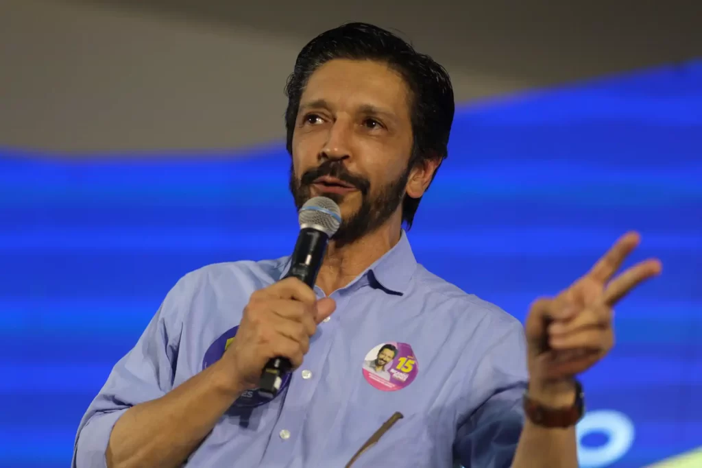 Homem em camisa azul com microfone e adesivo na lapela, gesticulando durante um discurso, destacando sua mensagem de campanha.