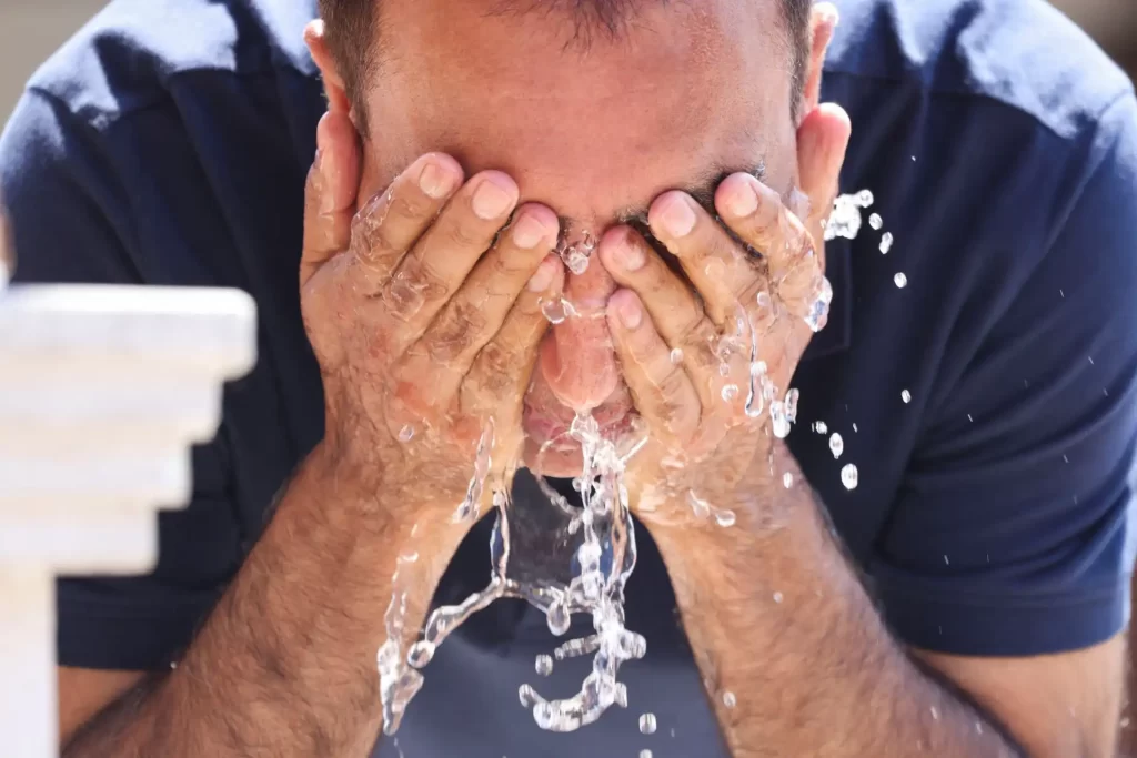 Homem lavando o rosto com água fresca em um dia ensolarado, promovendo a sensação de alívio e refresco. Ideal para temas de cuidado pessoal e bem-estar.