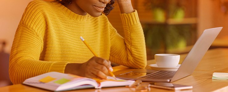 Mulher com suéter amarelo escreve em um caderno enquanto usa um laptop em um ambiente de trabalho aconchegante. A cena retrata foco e produtividade.