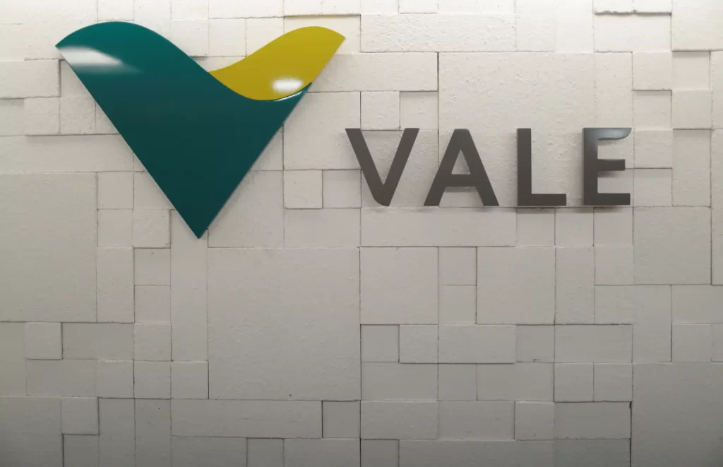 Logotipo da Vale em destaque, exibindo as cores verde e amarelo, simbolizando a identidade visual da empresa em uma parede de fundo neutro.