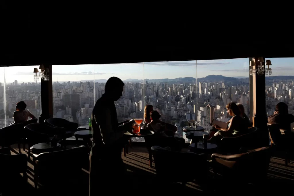 Vista deslumbrante de um lounge com pessoas desfrutando de drinks e refeições, em um ambiente sofisticado e iluminado, sobre uma cidade vibrante.