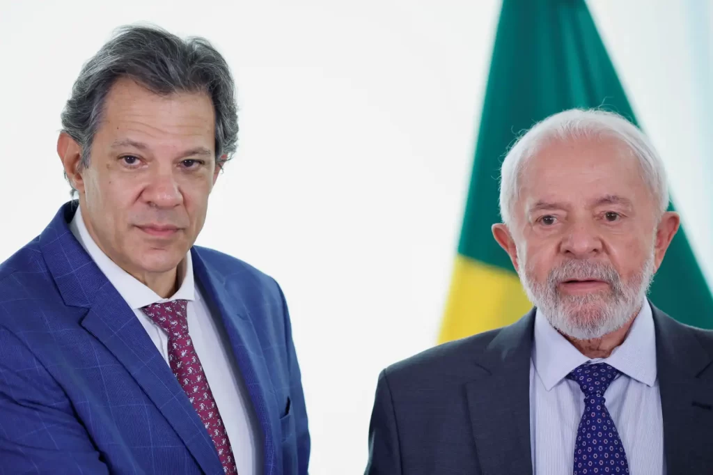 Lula e Fernando Haddad em um evento oficial, simbolizando a colaboração política e econômica no Brasil. Ambos vestem ternos formais e posam em frente à bandeira do Brasil.