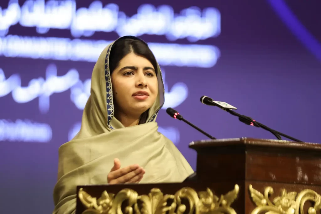 Malala Yousafzai se apresenta em um evento, falando sobre educação e direitos das mulheres, usando um vestido tradicional. Ela é uma ativista reconhecida mundialmente por sua luta.