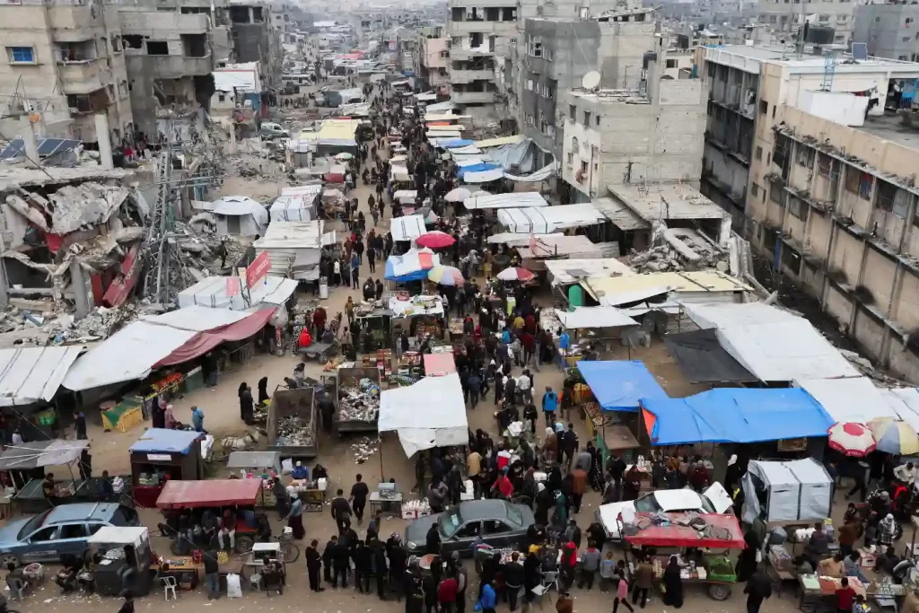 Vista aérea de uma movimentada rua de mercado em uma cidade em ruínas, com barracas coloridas e multidão de pessoas. A cena retrata a vida cotidiana em meio à destruição.