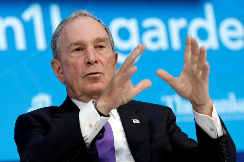 Michael Bloomberg em uma conferência, gesticulando durante seu discurso. Ele é conhecido por sua atuação na política e negócios.