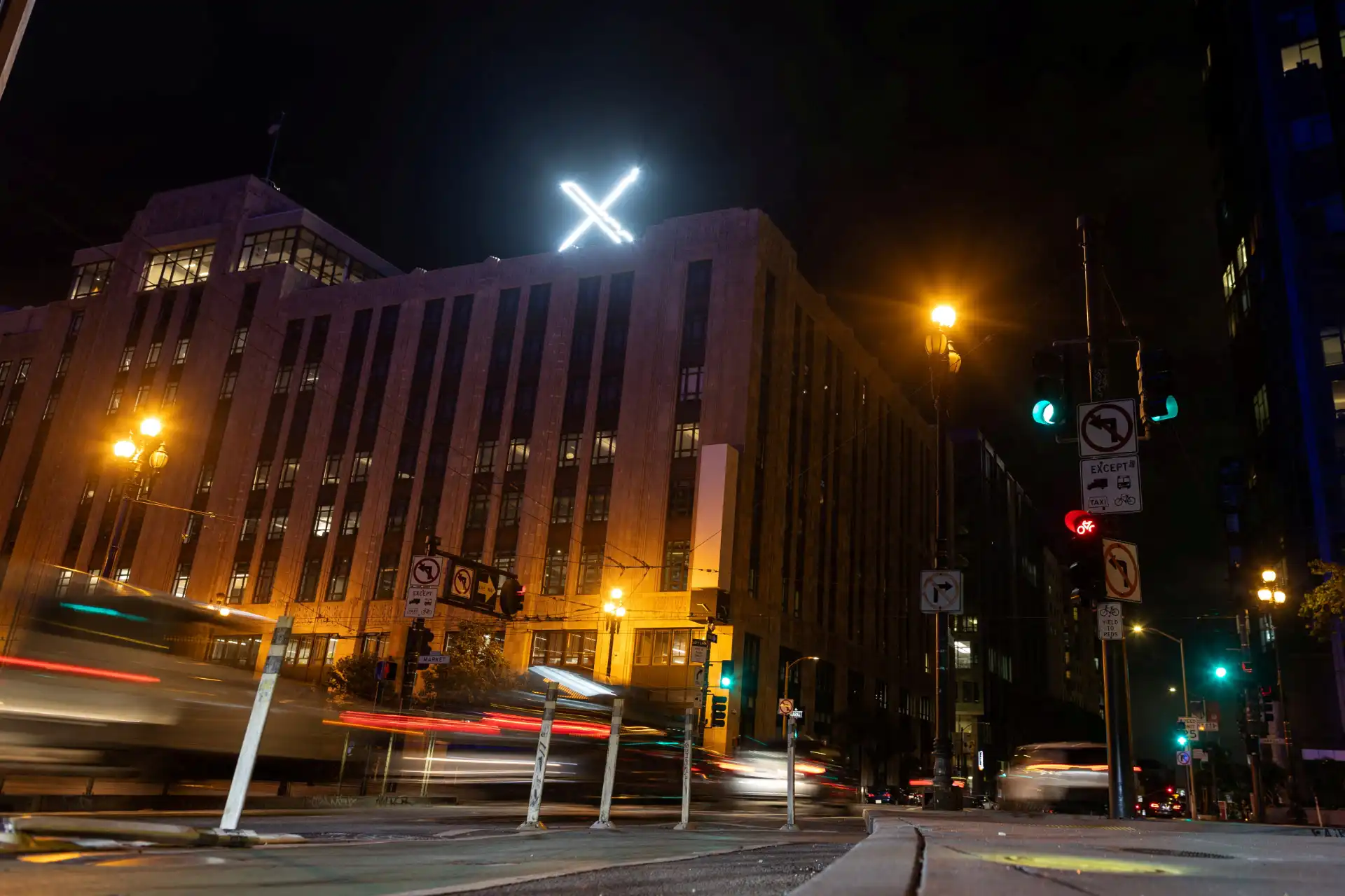 Paisagem urbana à noite com luzes de tráfego e um prédio iluminado com uma cruz no topo. Captura dinâmica de veículos em movimento.