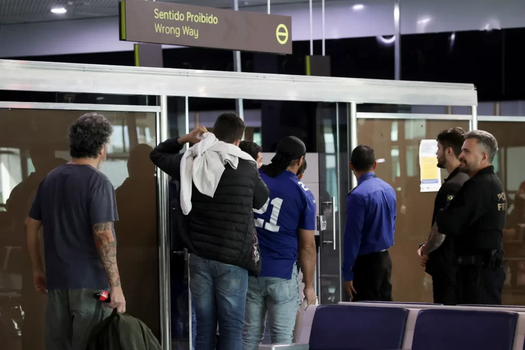 Passageiros em um aeroporto seguindo para a entrada errada com sinalização de 'Sentido Proibido' e presença de agentes de segurança.