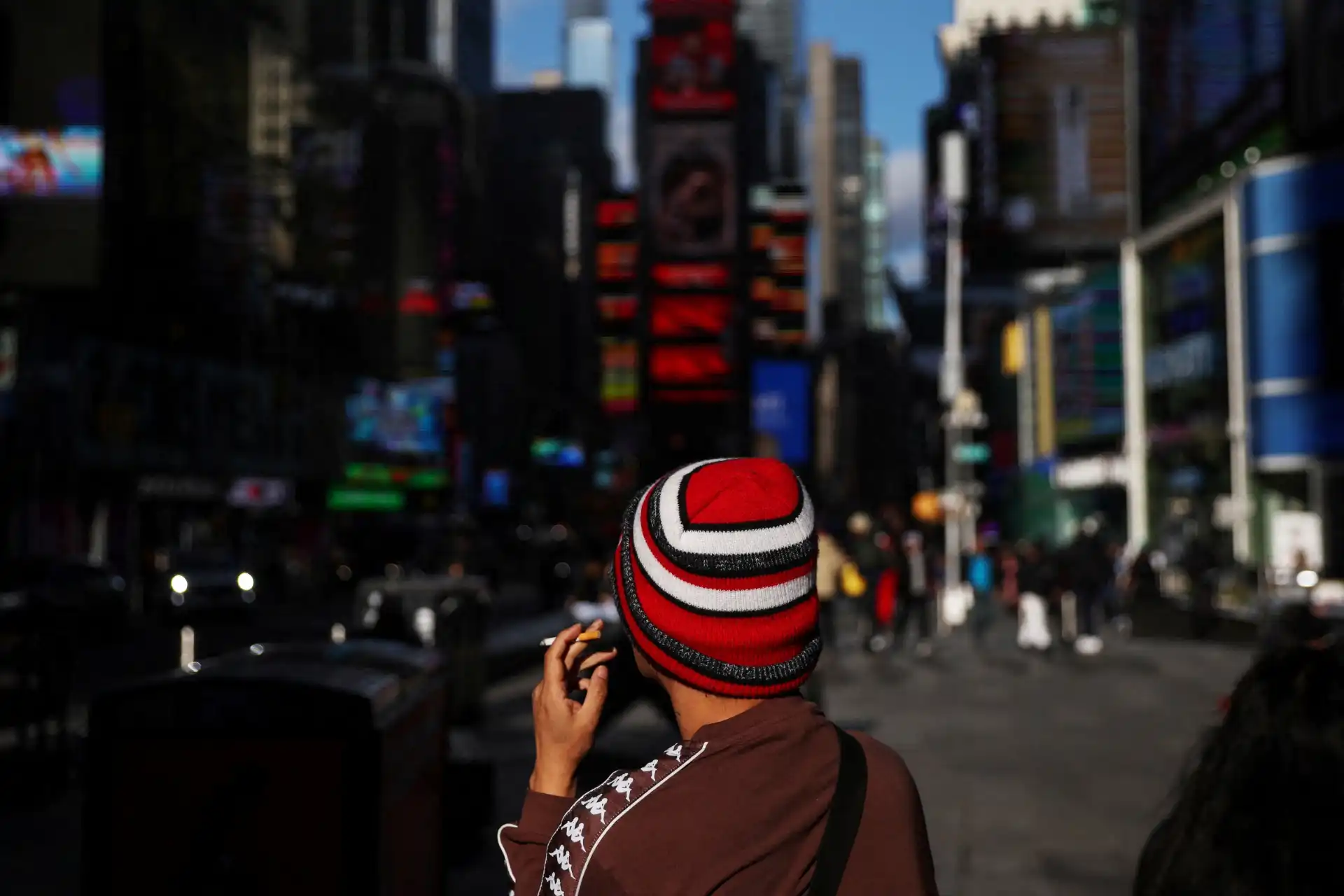 Pessoa em Times Square, Nova York, usando um gorro vermelho e preto, apreciando a vista da cidade moderna e vibrante, com letreiros luminosos ao fundo.