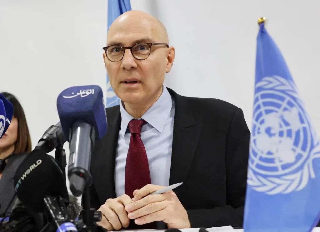 Imagem de um representante da ONU durante uma coletiva de imprensa, cercado por microfones e bandeiras da organização, discutindo temas relevantes.