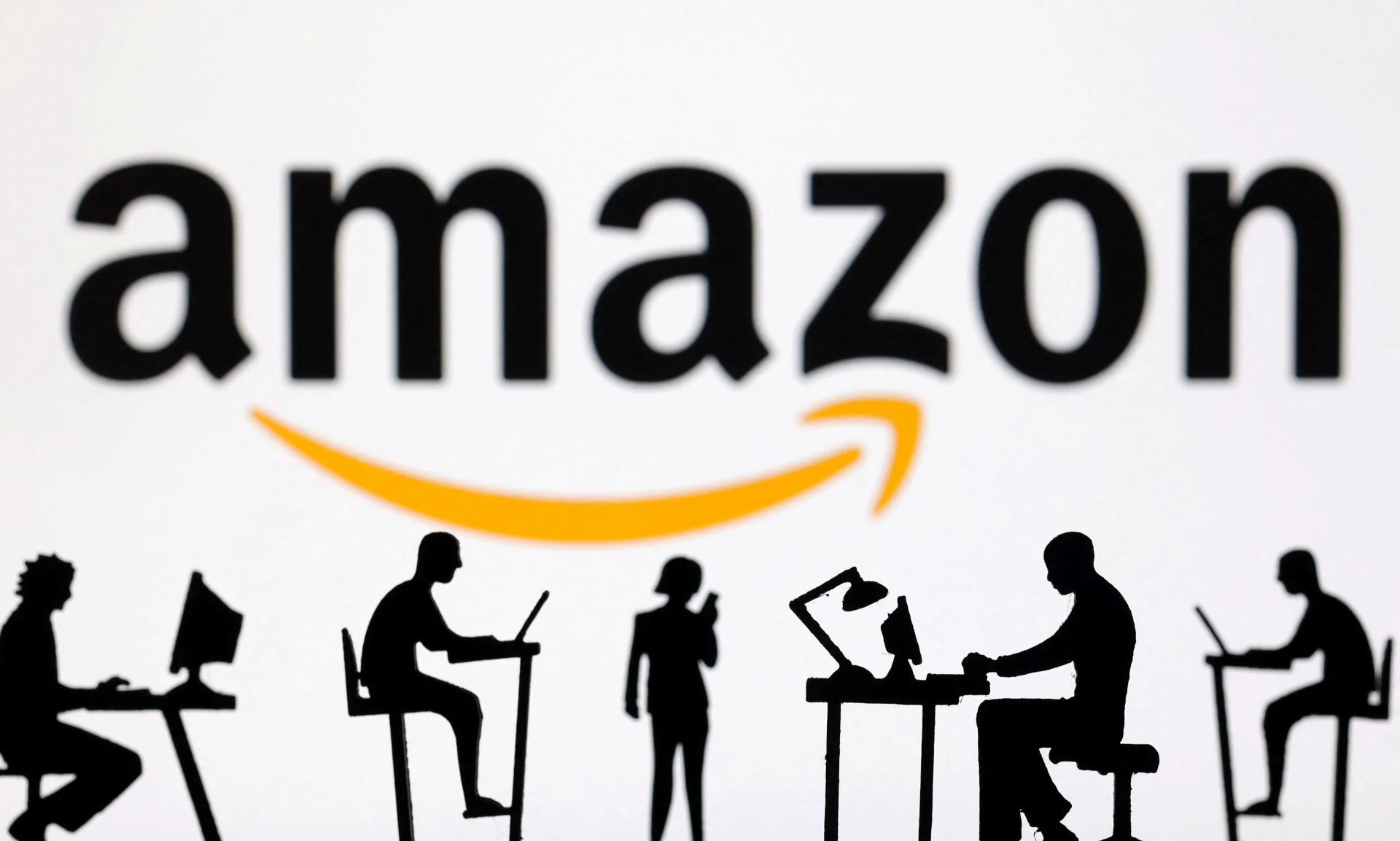 Silhuetas de pessoas trabalhando em computadores com o logotipo da Amazon ao fundo, representando o ambiente corporativo e a inovação tecnológica.