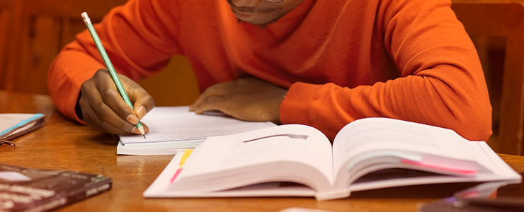 Menino estudando e escrevendo em um vade mecum com livros abertos em uma mesa de madeira, concentrado em suas atividades escolares.