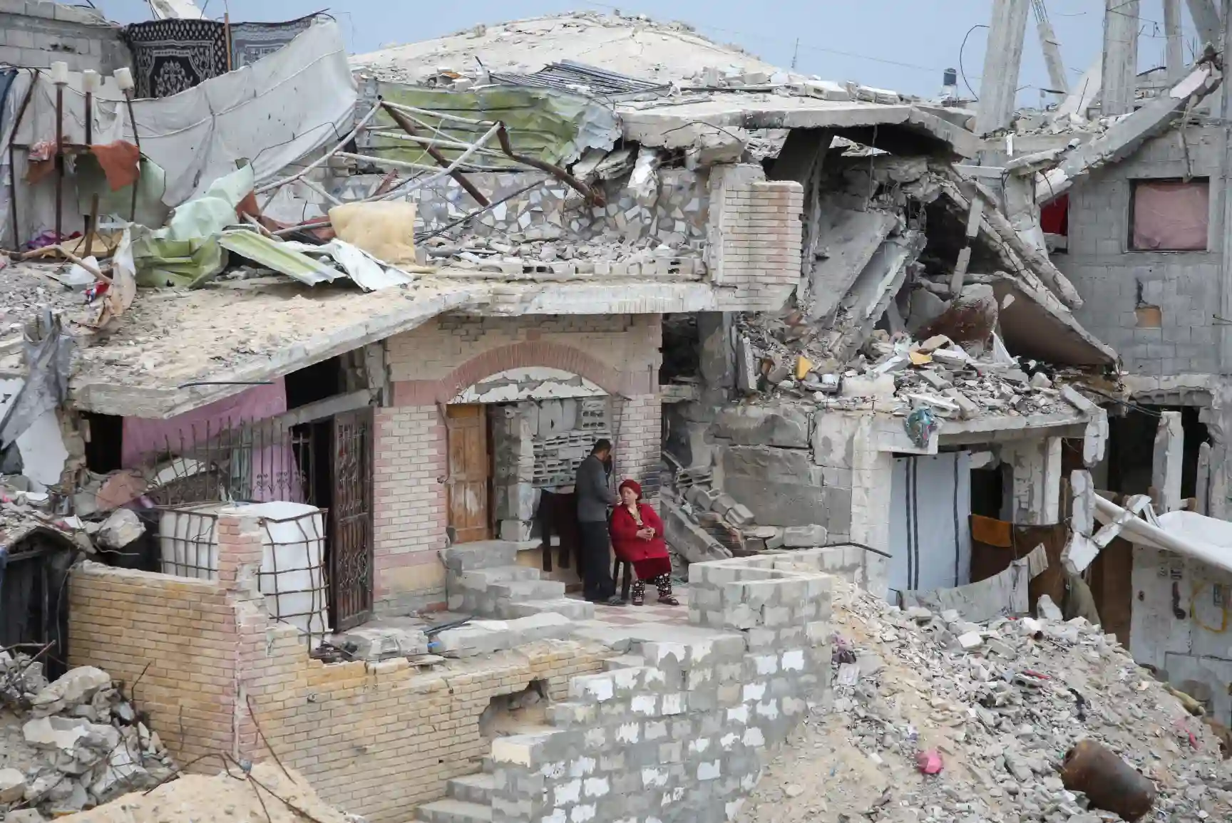 Imagem de uma área residencial devastada, mostrando edifícios em ruínas e pessoas sentadas nas escadas, refletindo os efeitos de um desastre.