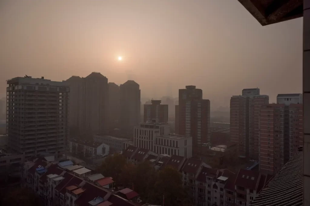 China pretende eliminar poluição atmosférica severa este ano Vista de uma cidade envolta em poluição atmosférica, com o sol nascendo entre prédios altos. A imagem retrata a realidade da qualidade do ar nas áreas urbanas, destacando a necessidade de cuidar do meio ambiente e melhorar a qualidade do ar nas cidades.