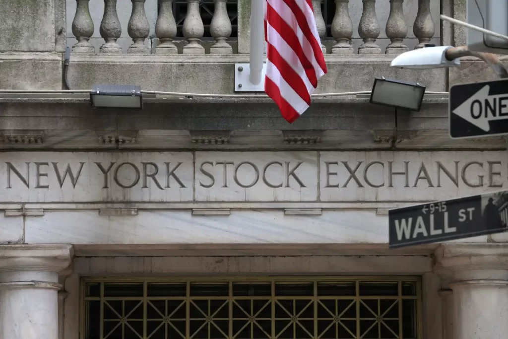 Entrada da Bolsa de Valores de Nova York com a bandeira dos EUA e placas de sinalização indicando Wall Street. Local histórico para investimentos.