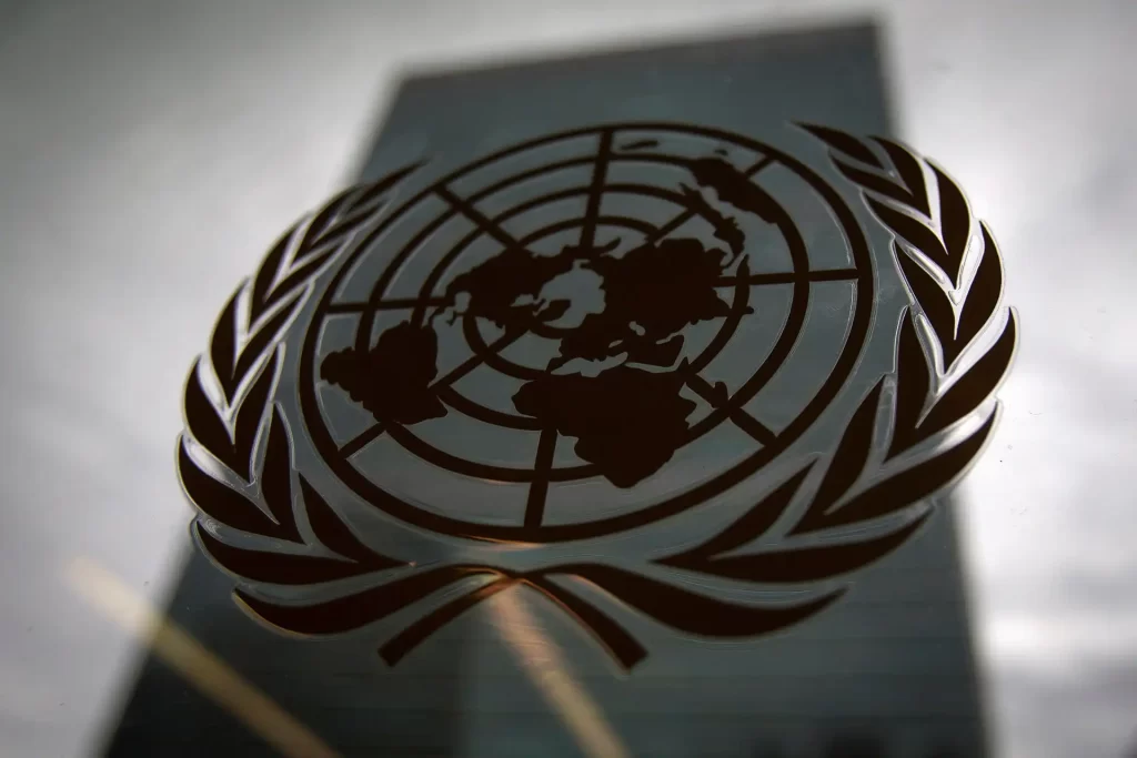 Logotipo da ONU em primeiro plano, com o fundo de um edifício refletido, simbolizando a paz e a colaboração internacional.