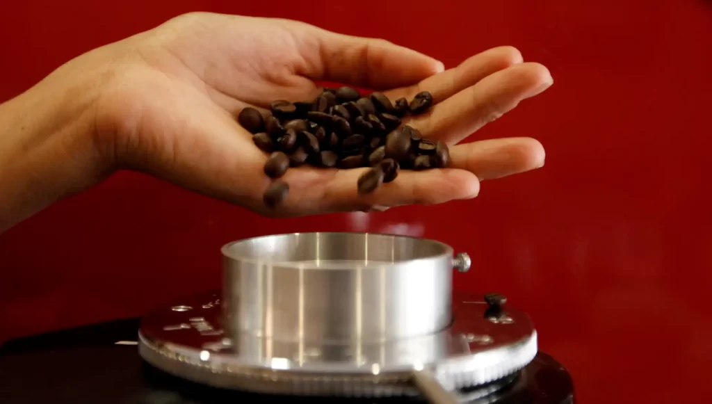 Mão segurando grãos de café em frente a uma balança, representando a moagem e preparo do café fresco.