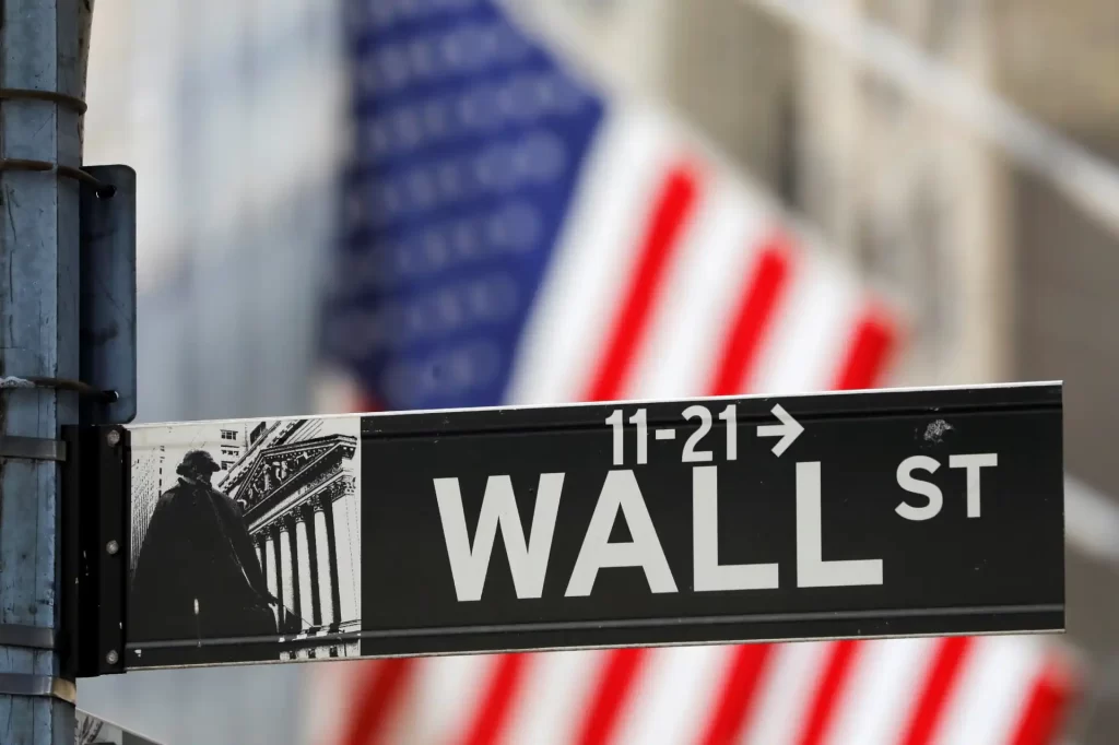 Placa da rua Wall Street, em Nova York, conhecida como o centro financeiro do mundo, com bandeira americana ao fundo.