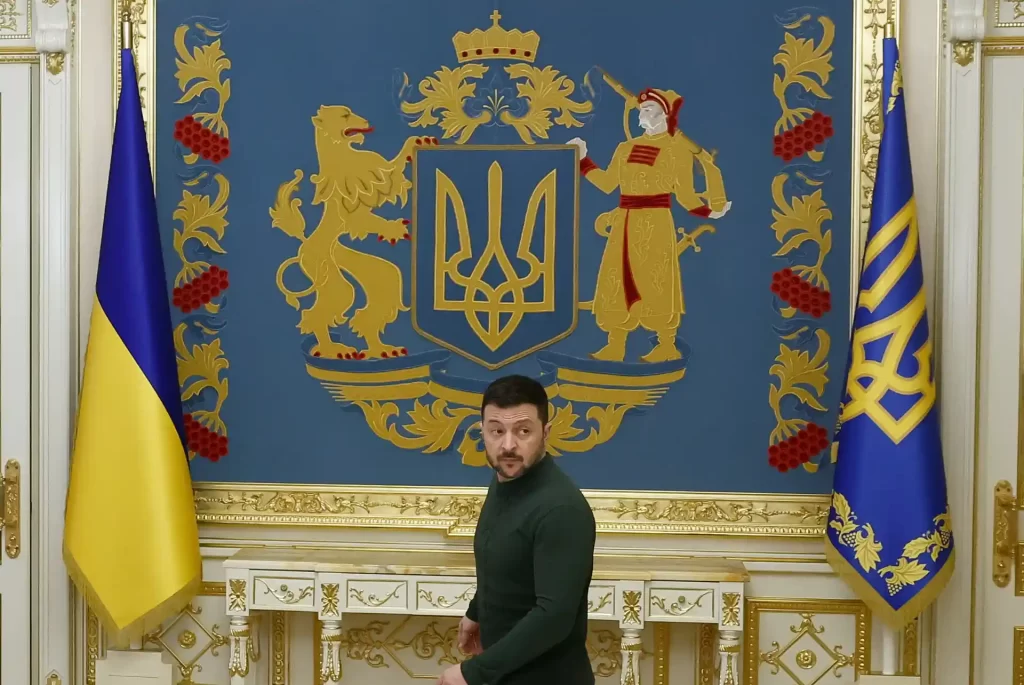 Imagem do presidente da Ucrânia, Volodymyr Zelensky, posando em frente ao brasão e bandeiras da Ucrânia, simbolizando a soberania e a cultura do país.
