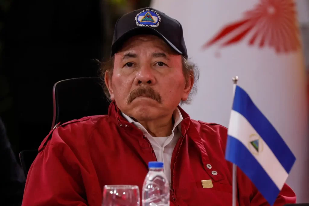 Retrato de um homem com um boné e uma jaqueta vermelha, observando atentamente enquanto uma bandeira da Nicarágua está à sua frente. A cena transmite um olhar sério e focado.