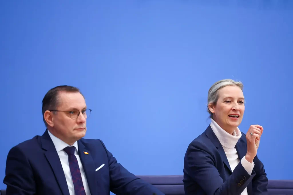 Imagem de uma reunião com dois indivíduos em primeiro plano, um deles gesticulando enquanto fala, com um fundo azul. Eles discutem temas importantes.