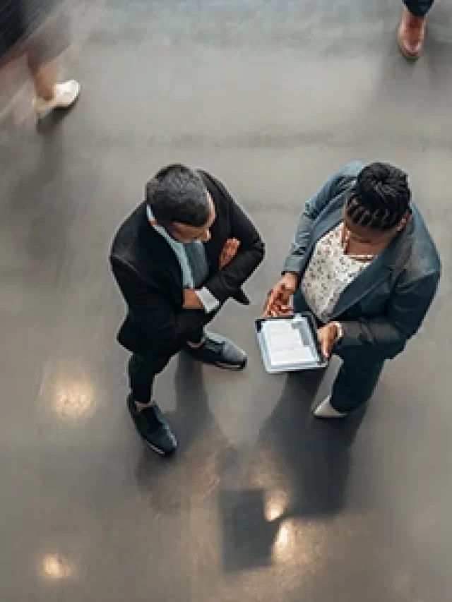 Story Imagem de pessoas em um ambiente corporativo, duas pessoas conversando e utilizando um tablet. Movimento ao fundo indica dinâmica entre os trabalhadores.