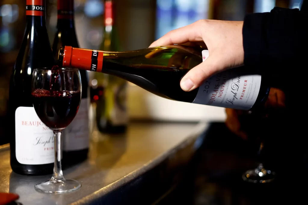 Uma mão derramando vinho tinto de uma garrafa em uma taça em um bar, com garrafas de vinho ao fundo. Perfeito para quem aprecia uma boa bebida.