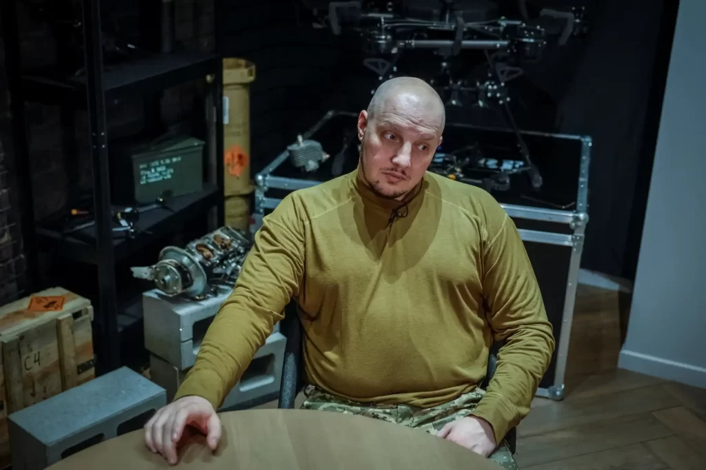 Homem sentado à mesa em ambiente com equipamentos tecnológicos, vestindo roupa camuflada e camiseta de manga longa, parece pensativo e atento.