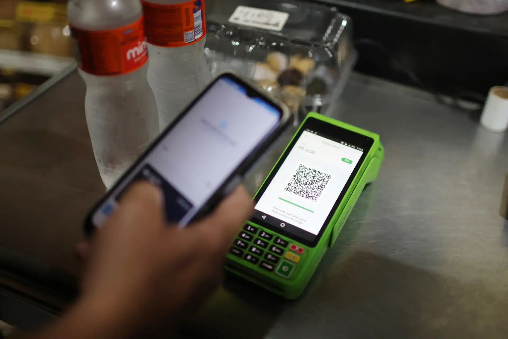 Pessoa utilizando smartphone para realizar pagamento com QR Code em um terminal verde. Garrafas de água e doces ao fundo.