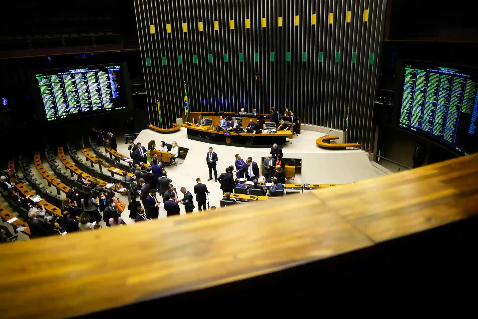 Plenário da Câmara dos Deputados durante uma sessão com representantes em discussão e painéis de votação visíveis. O ambiente é caracterizado por uma arquitetura moderna.