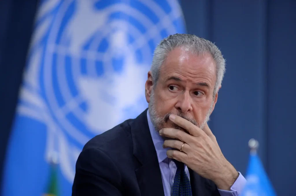 Imagem de um representante da ONU em uma conferência, refletindo sobre questões globais. A bandeira da ONU e do Brasil em segundo plano.