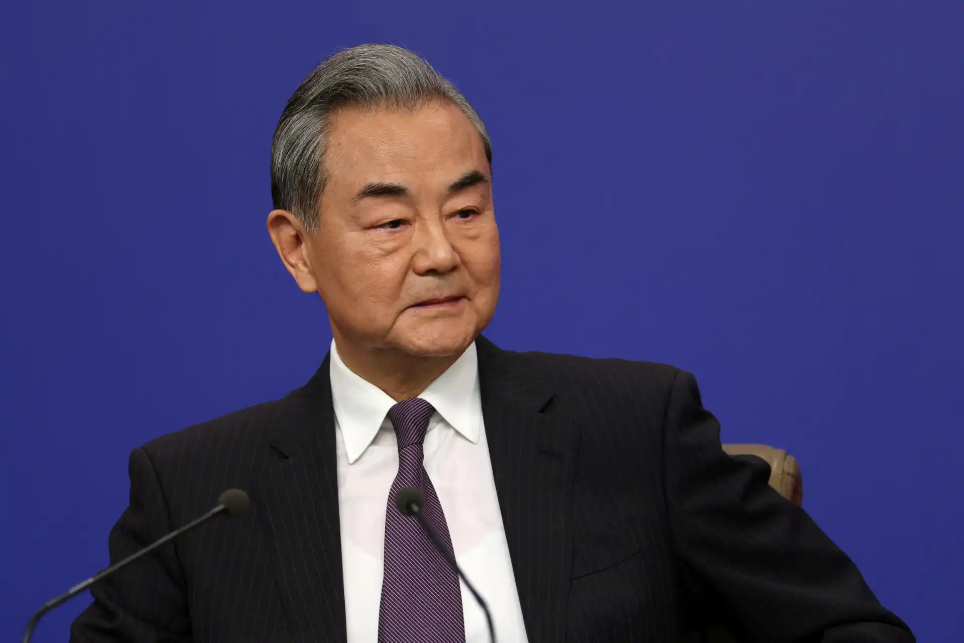 Wang Yi, diplomata chinês, em um momento de conferência, com expressão serena e vestindo terno escuro, destacado em fundo azul.