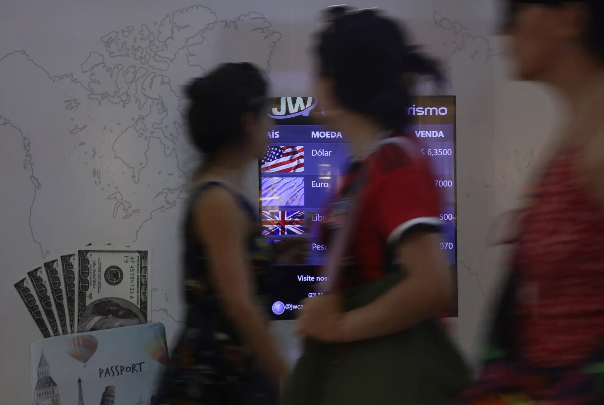 Imagem de um local de câmbio com várias pessoas passando, mostrando cédulas de dólar e uma tela com as taxas de câmbio para várias moedas.
